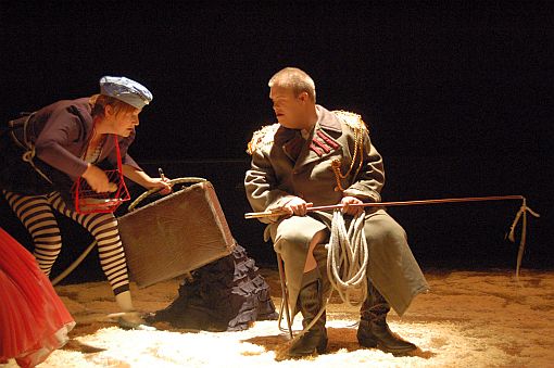 Foto: Szene aus dem Stück "Godot lässt grüßen" - Schauspieler mit Peitsche in Uniform und Schauspielerin mit Koffer in der Hand, die tanzt